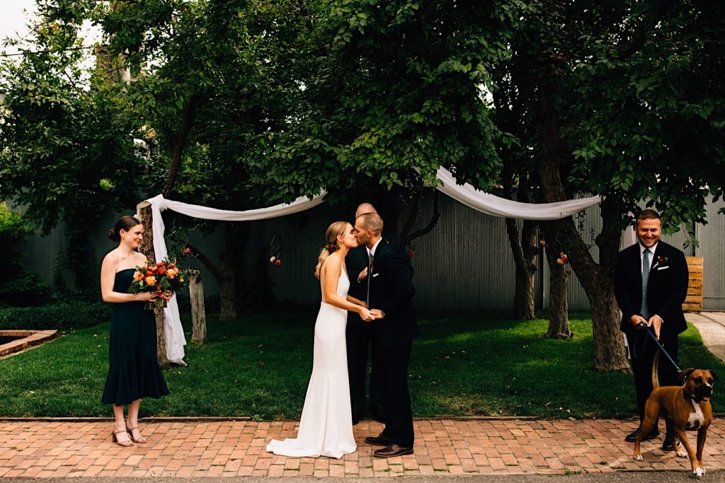 A bride and groom kiss during their intimate outdoor wedding ceremony, there are trees behind them, the best man and the couples dog stand on the right while the maid of honor stands on the left holding a bouquet of flowers
