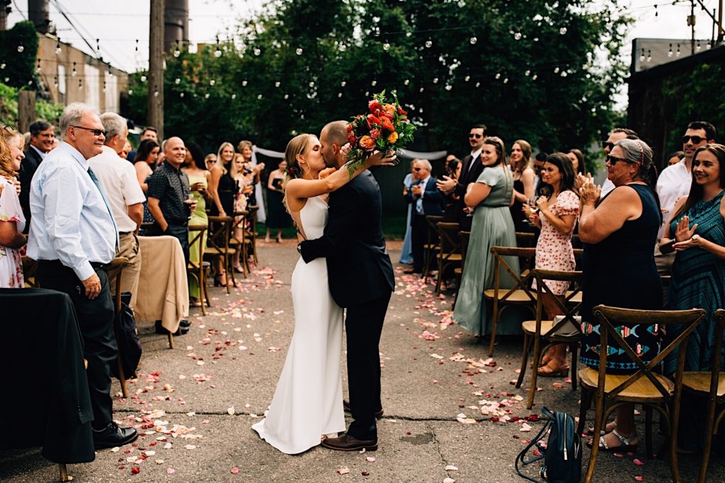 A bride and groom kiss after their intimate outdoor wedding reception at The Ramble Hotel in downtown Denver, the bride holds a flower bouquet behind the grooms head as their guests stand and clap on either side of the couple