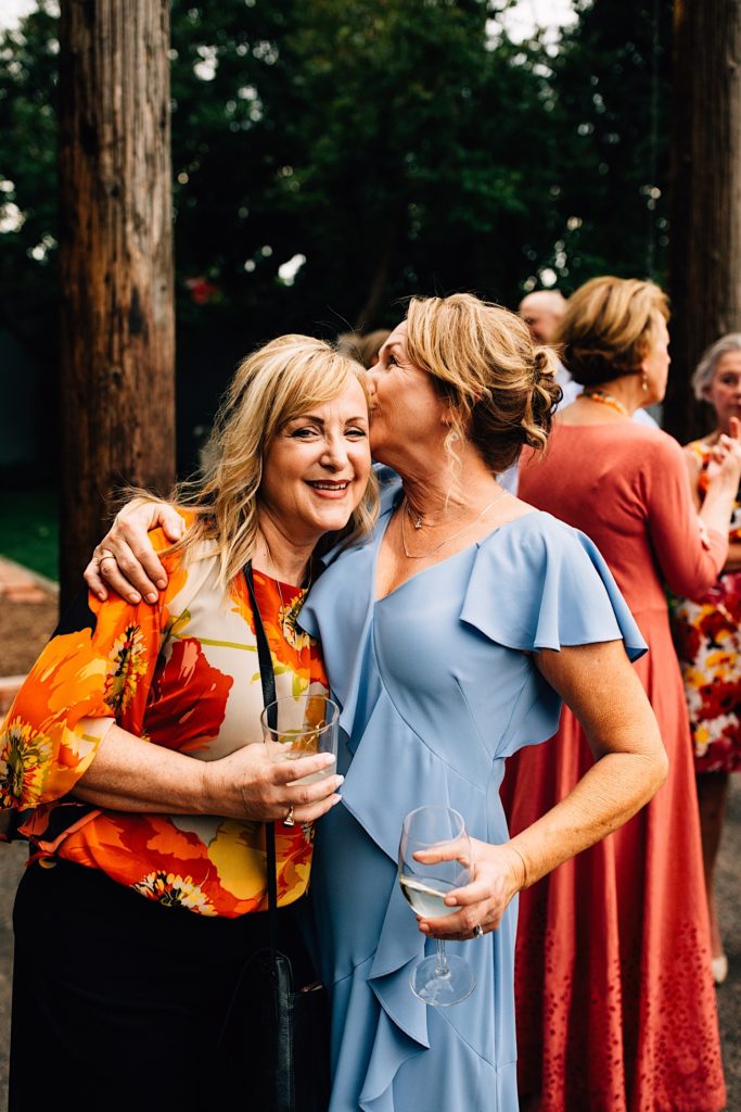 Two woman standing next to one another during a wedding with other guests behind them, the woman on the left smiles at the camera while the woman on the right kisses her temple, both are holding drinks