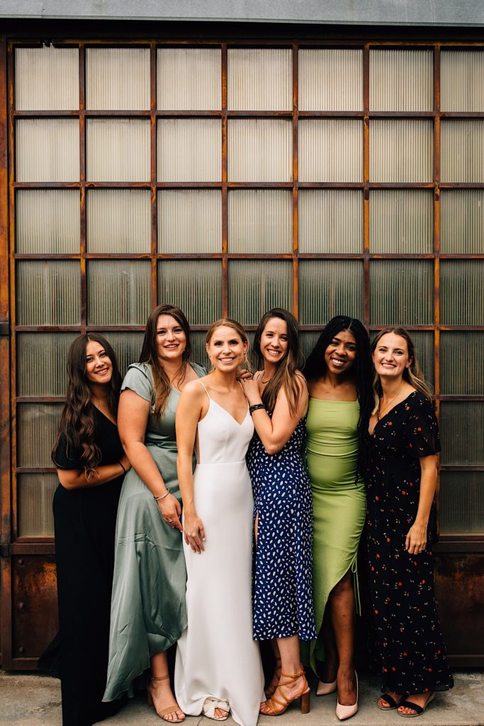 A bride stands with her friends on either side of her as they all smile at the camera with a wood and glass wall behind them