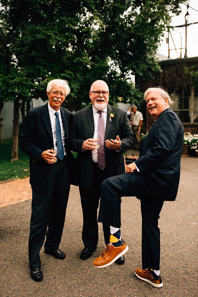 Three men wearing suits stand together and smile at the camera during a wedding cocktail hour, the man on the right lifts one leg to show off his brown shoes and checkered socks