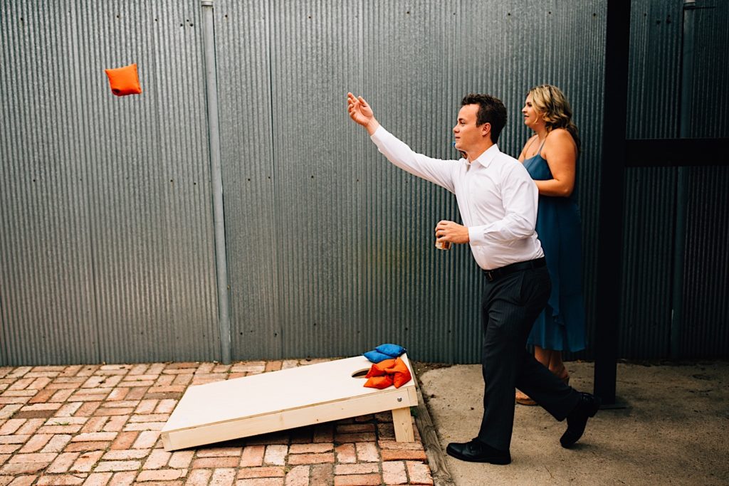 A man and a woman playing cornhole at a wedding reception at the Ramble Hotel, the man has just tossed a red bean bag and is holding a beer