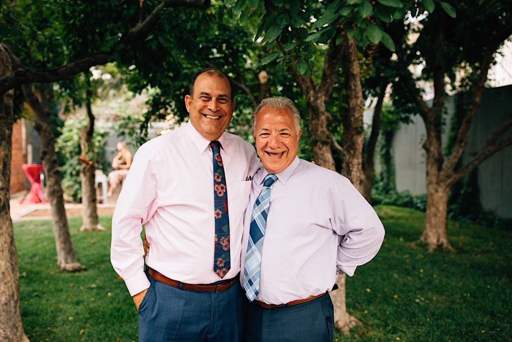 Two men standing next to one another smiling at the camera, they're dressed up for a wedding and standing underneath trees