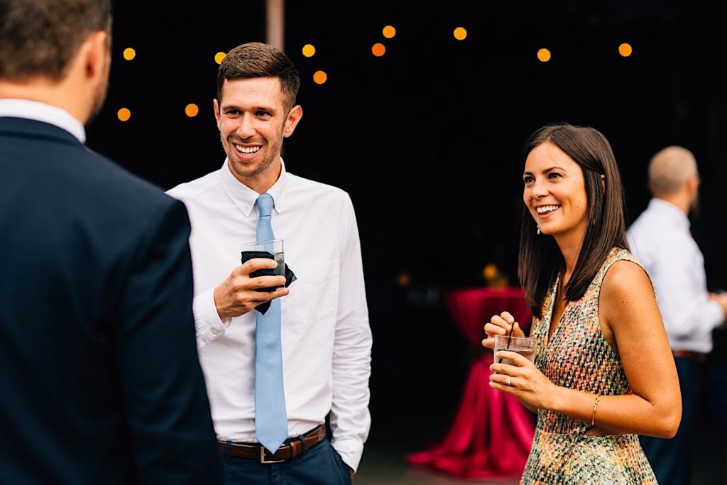 A man and a woman holding drinks chat with a man that has his back to the camera during a wedding reception