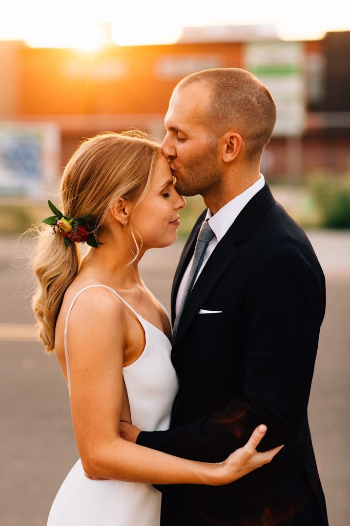 Groom kisses his bride's forehead while wearing their wedding attire and the sun sets behind them