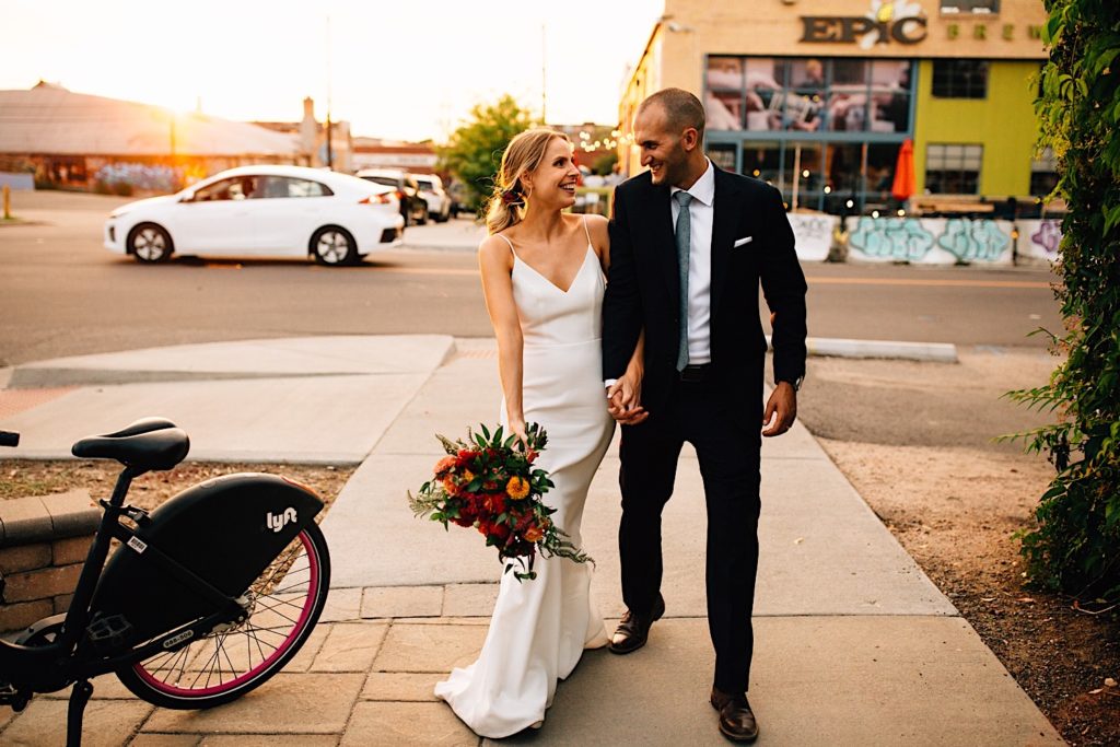 A bride and groom in wedding attire walk towards the camera and smile at one another while holding hands, the sun is setting behind them in downtown Denver 