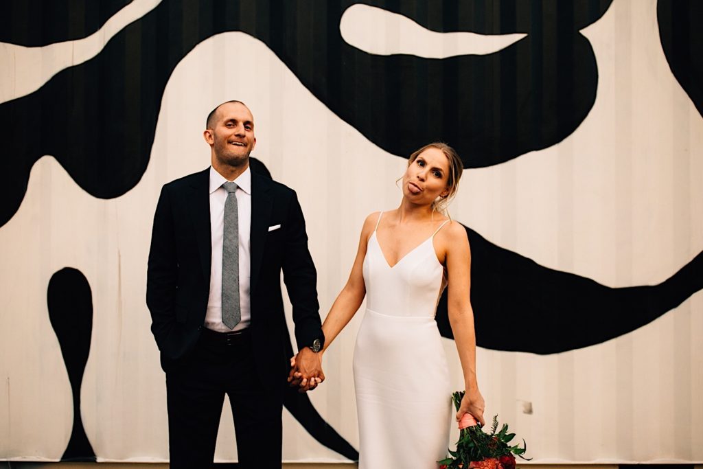 Bride and groom standing next to one another holding hands and looking at the camera making funny faces, the bride is holding a flower bouquet and they are in front of metal siding with a clack and white cow pattern on it