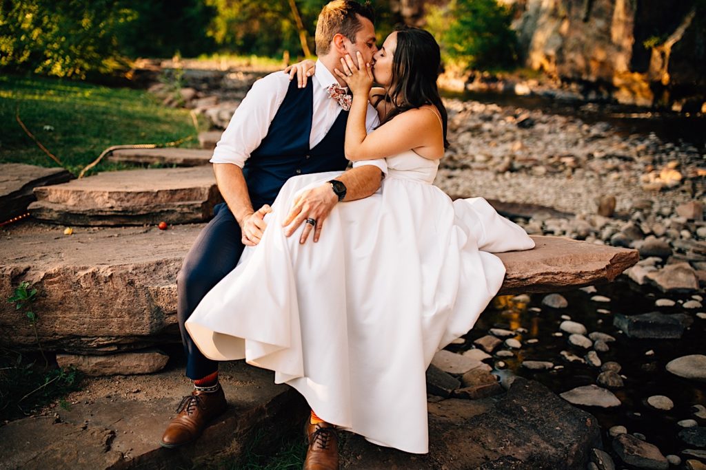 Bride and groom sit next to each other on a stone and kiss at Planet Bluegrass