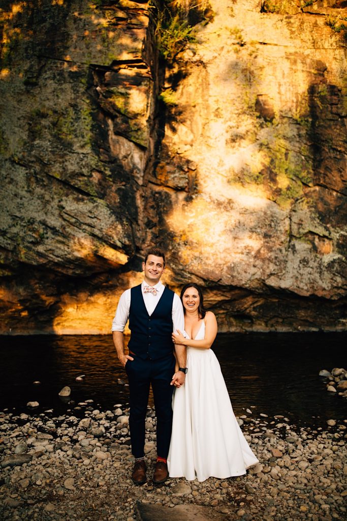 Bride and groom hold hands and stand next to each other in front of a rock wall and river at Planet Bluegrass