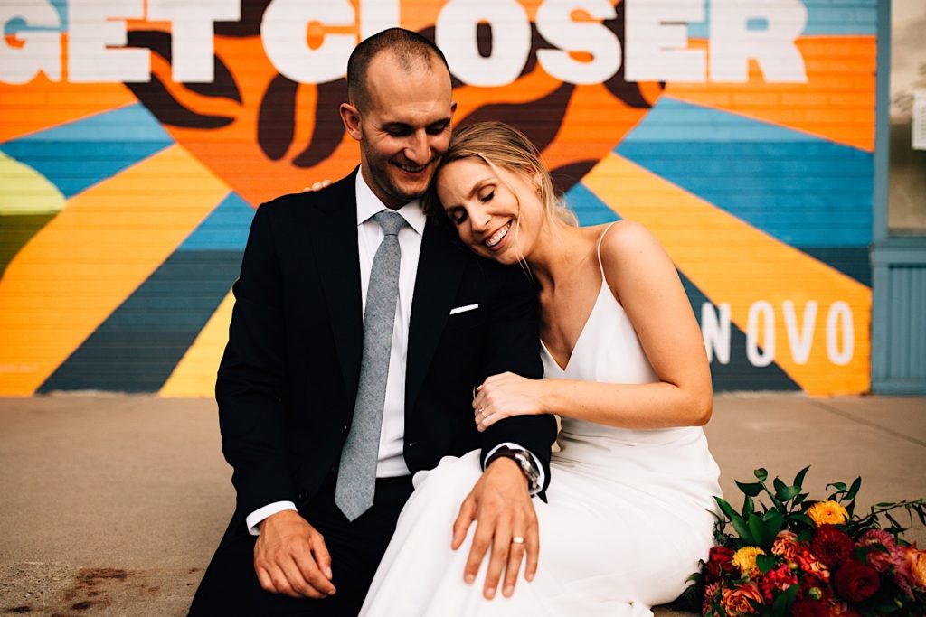 A bride and groom sit on the sidewalk together smiling while wearing their wedding attire in downtown Denver, there is a mural behind them that reads " Get Closer, Novo"