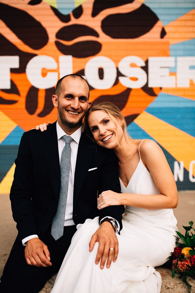 A bride and groom sit on the sidewalk together smiling at the camera while wearing their wedding attire in downtown Denver, there is a mural behind them that reads "Closer"