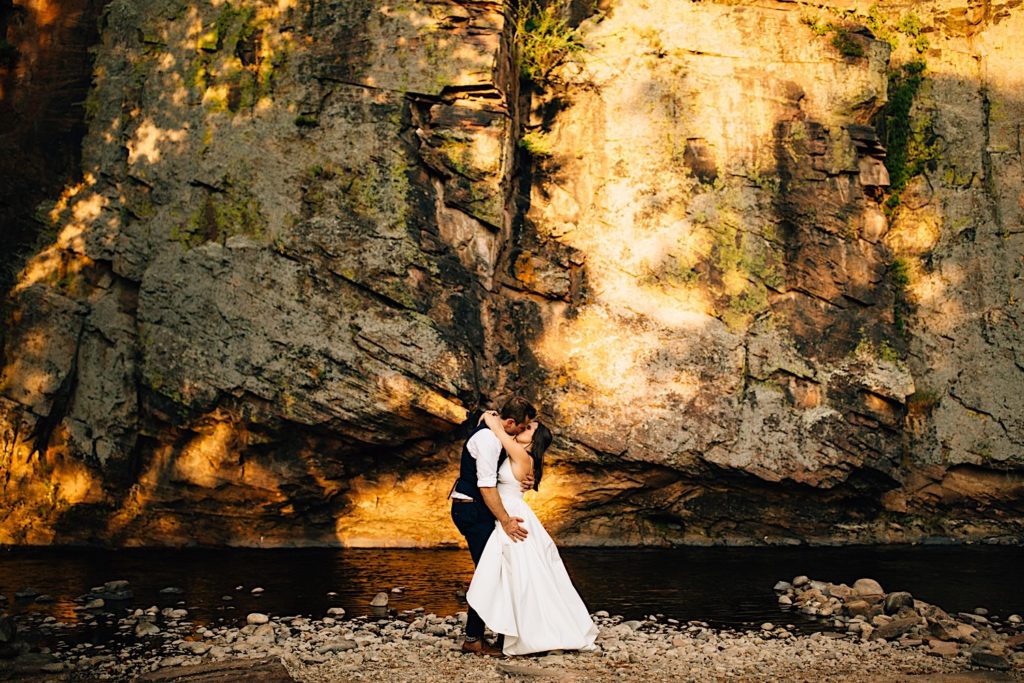 Bride and groom kiss in front of a rock wall and river at Planet Bluegrass