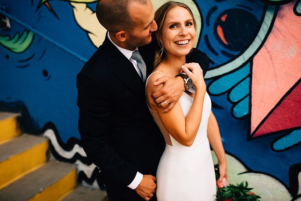 A groom stands behind a bride with his arm wrapped around her, the groom smiles at the bride while she smiles at the camera, they're wearing their wedding attire and there is a colorful mural in downtown Denver behind them