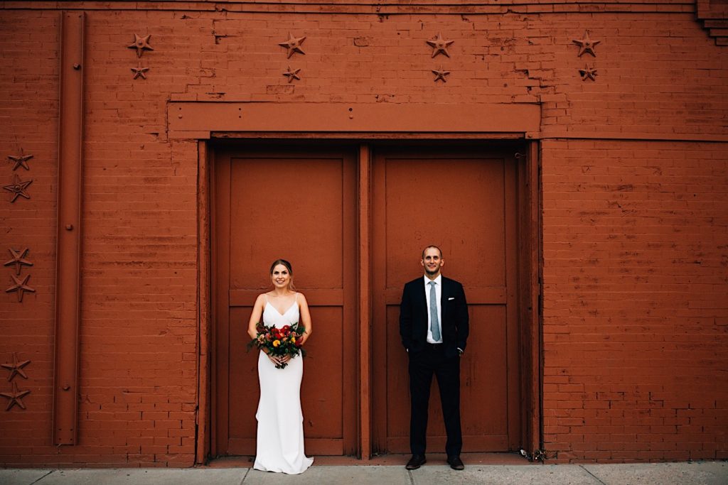 Bride and groom standing in their wedding attire in front of a orange/brown brick wall smiling at the camera, there is a double doorway on the wall and they are each in front of one door