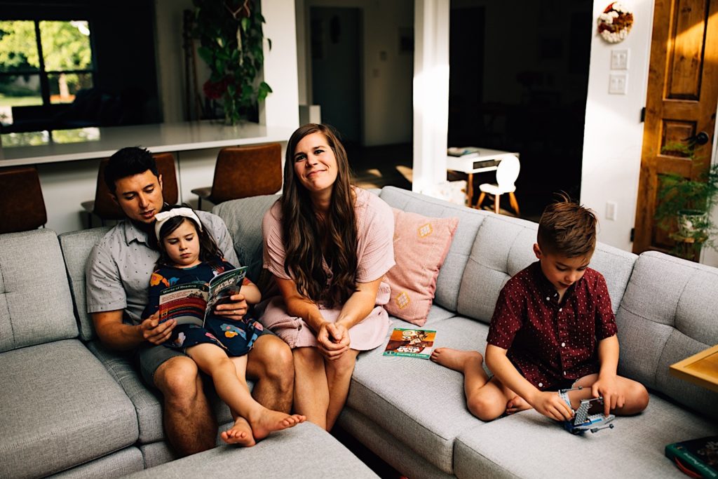 A family sits together on the couch, the father helps the daughter read while the mother smiles at the camera as her son plays with Legos next to her