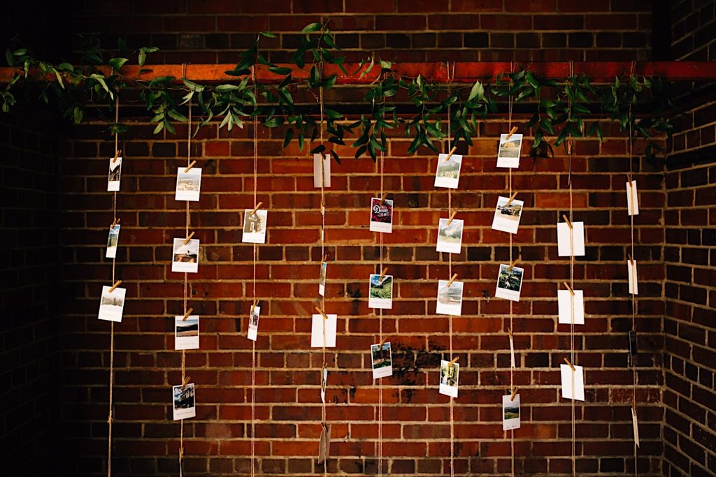 Polaroids clipped to pieces of string dangle from the ceiling in front of a brick wall