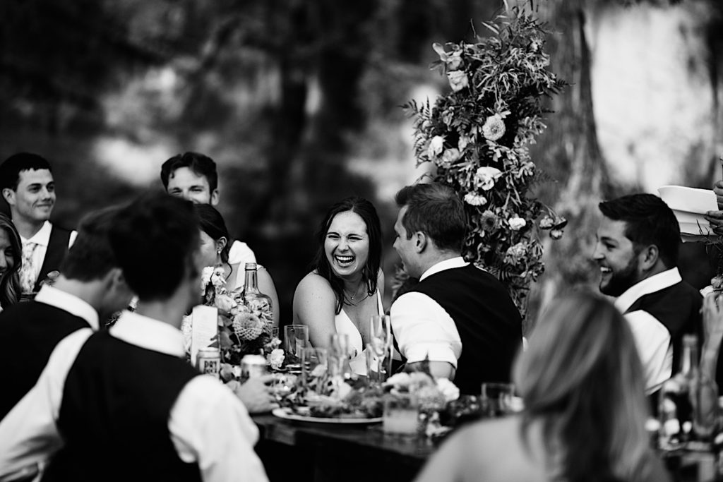 Black and white photo of a bride laughing surrounded by her wedding party and seated next to the groom