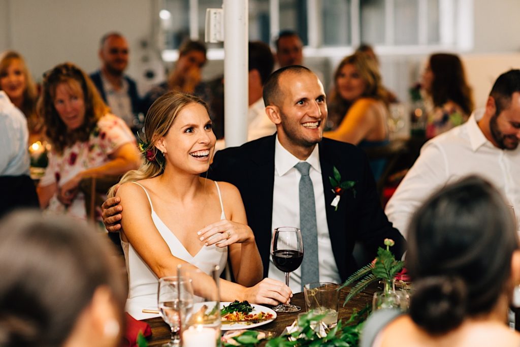 Bride and groom sitting next to one another at a table during their wedding reception and smiling, the groom has his arm around his bride and they are surrounded by other seated guests