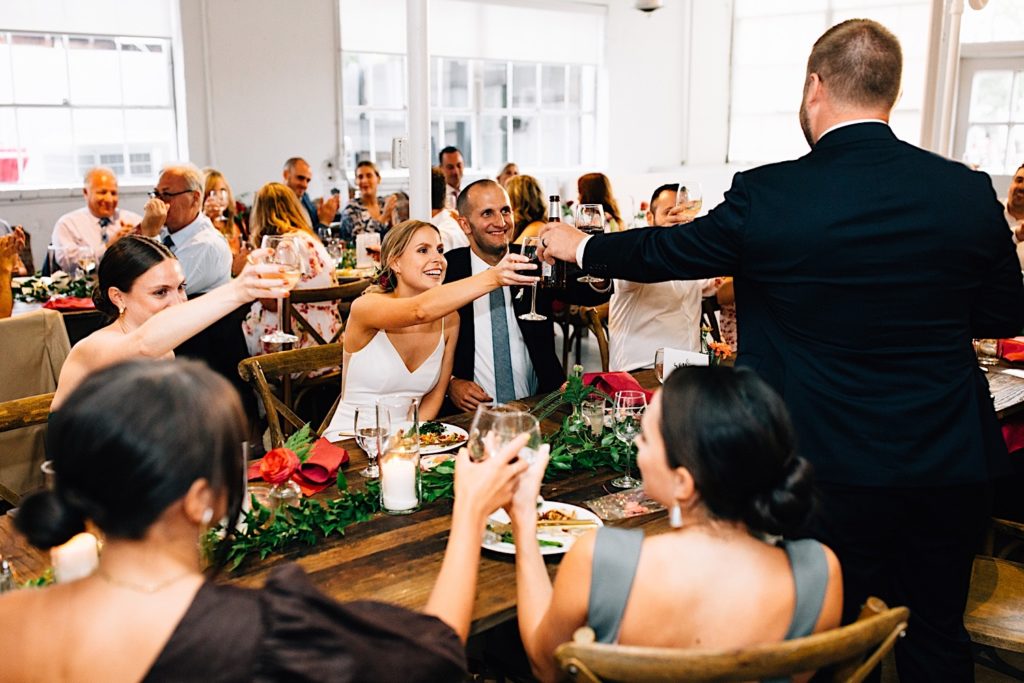 A bride and groom sitting next to one another during their wedding reception toast the best man who has his back to the camera, guests around them also toast one another