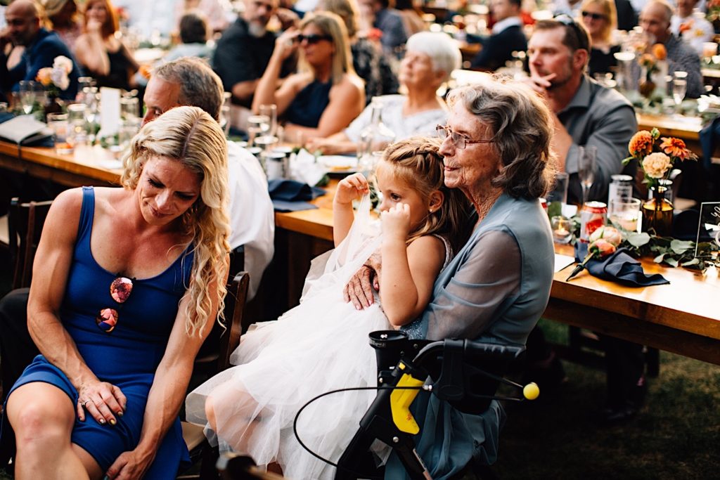 A grandma holds her granddaughter while seated with other guests during a wedding reception