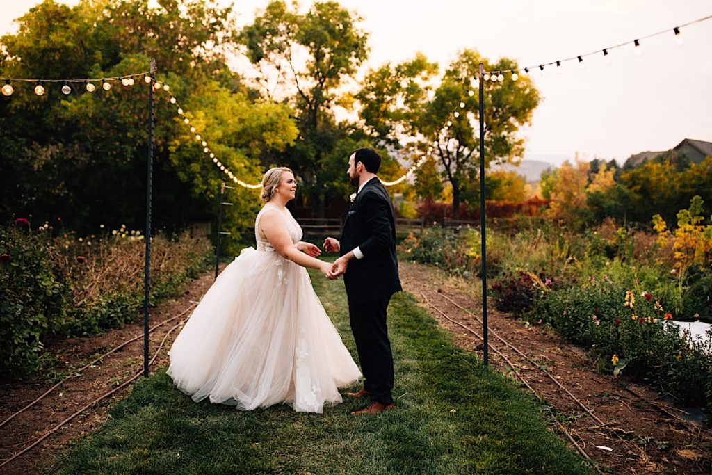 A bride and groom stand facing one another in their wedding attire in a garden in Colorado with string lights on either side of them.