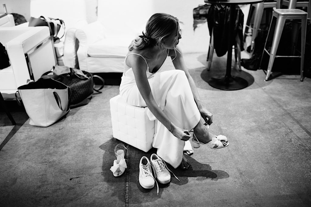 Black and white photo of a bride sitting and taking off her formal wedding shoes and putting on street shoes