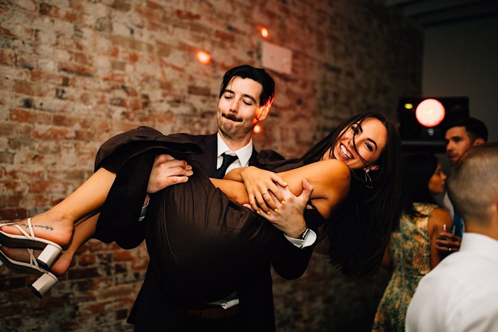A man with a mustache holds a woman up as she smiles at the camera while they're at a wedding reception