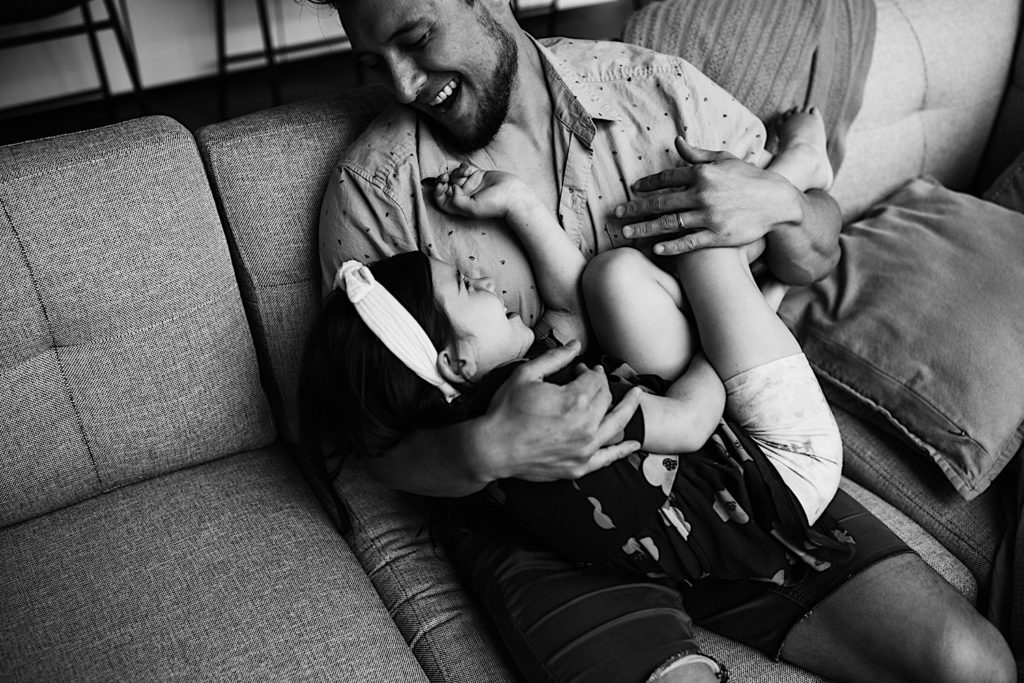 Black and white photo of a father smiling and laughing while holding his laughing daughter on the couch