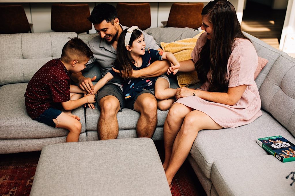 A family sits together on the couch smiling and laughing with each other, the son is holding his sisters arm while she's being held by the father.