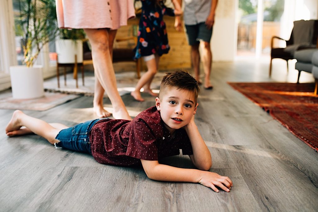 A child poses laying on the living room floor and looking at the camera while his family is standing behind him with only their legs visible