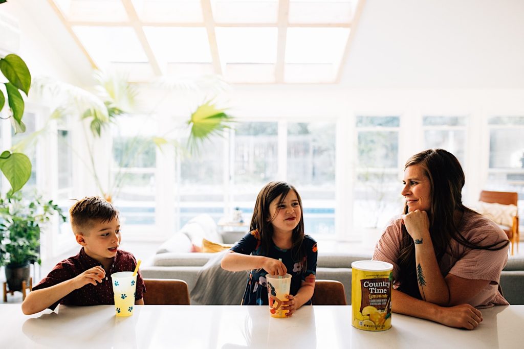 A family sits at a countertop together, the children have lemonade glasses in front of them while the mother smiles at them with the lemonade mix in front of her.