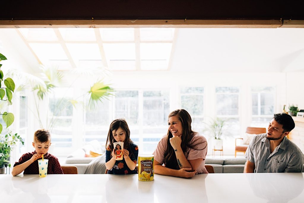 A family sits at a countertop together, the children have lemonade glasses in front of them while the mother and father smile at them with the lemonade mix in front of the mother.