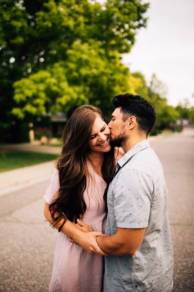 A couple embrace in the street, the woman smiles as the man kisses her on the cheek.