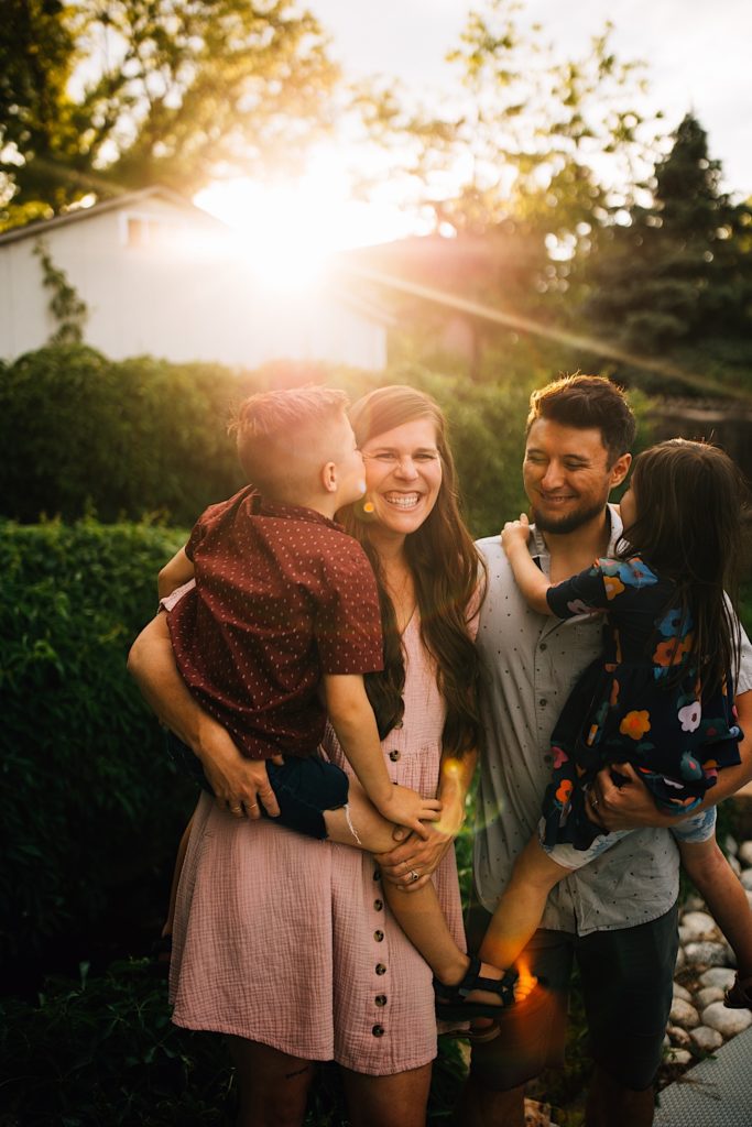 A family stand together in their backyard and smile as the sun sets, the mother is holding her son as he kisses her on the cheek while her husband holds their daughter next to her