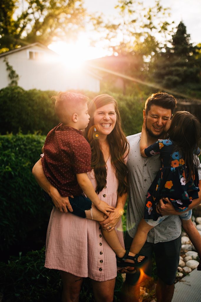 A family stand together in their backyard and smile as the sun sets, the mother is holding her son while her husband holds their daughter as she kisses his cheek