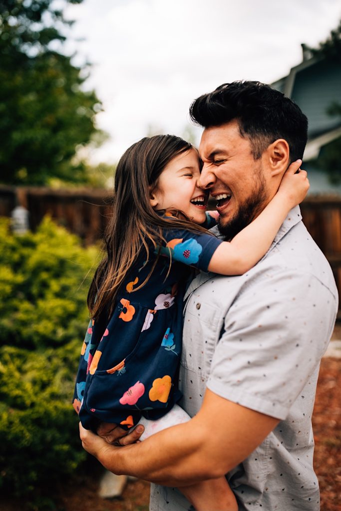 A father holds his daughter as they both laugh with their faces together and their eyes closed