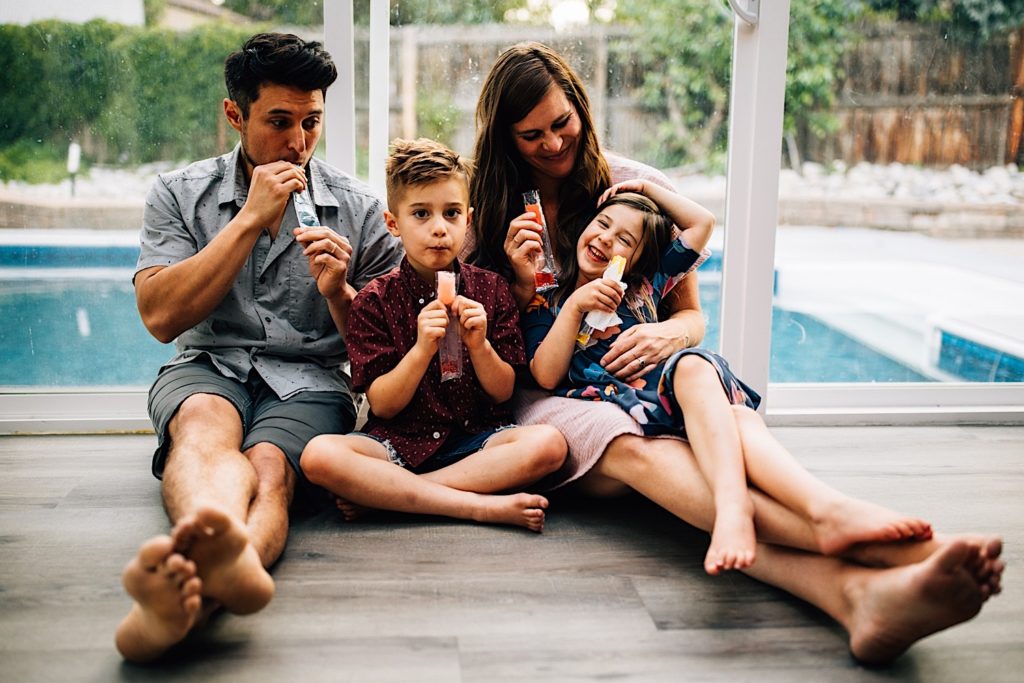 A family sits inside on the floor together eating freeze pops with their backs against a glass door with a pool behind them.