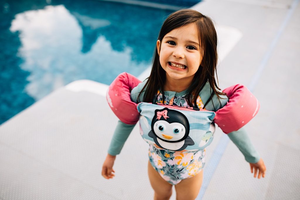 A child wearing a swimsuit and floaties stands next to a pool and smiles up at the camera