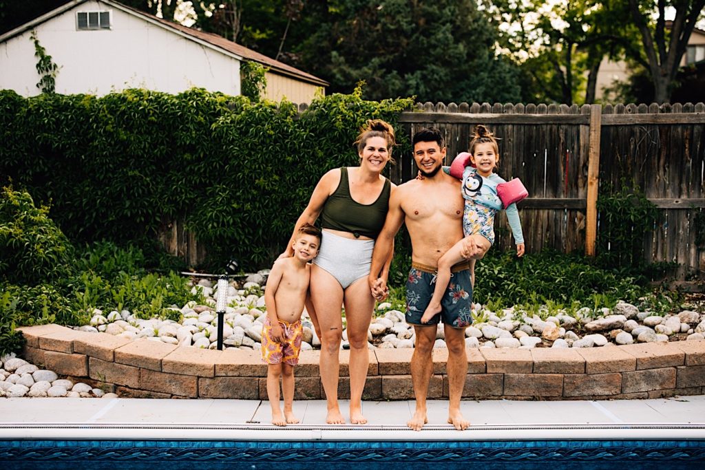 A family wearing their swimsuits stand outside next to their pool and smile at the camera. The father is holding his daughter while the mother stands next to them and their son