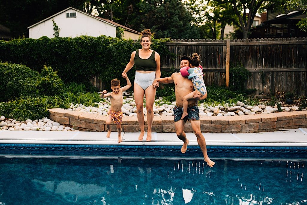 A family jump in the pool while all holding hands, the son is looking down at the pool, the mother smiles at the camera, and the father is holding the daughter with his eyes closed