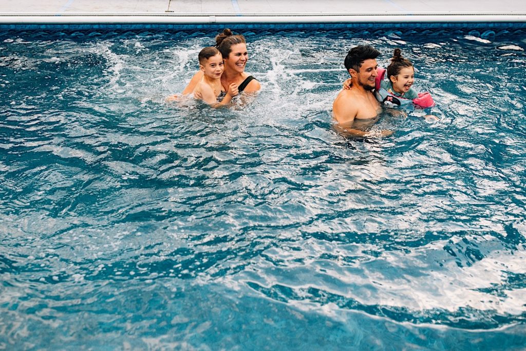 A family swim in their pool together, the mother holding her son as her husband holds their daughter next to them, they're all smiling and laughing together
