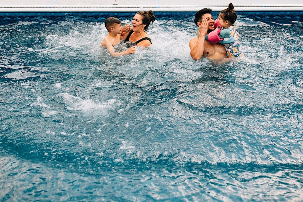 A family swim in their pool together, the mother holding her son as her husband holds their daughter next to them, they're all smiling and laughing together