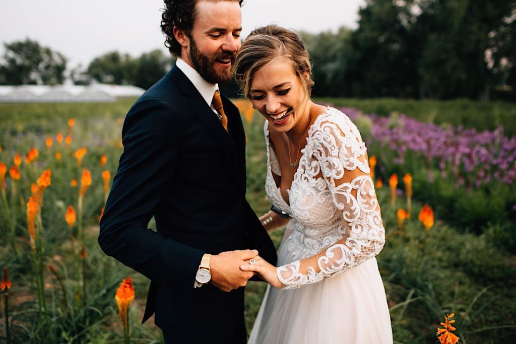 A bride and groom stand facing one another holding hands at Boulder Flower Farm. They're in their wedding attire and the bride is laughing as the groom smiles