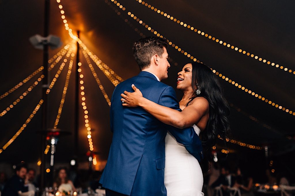 A bride and groom share their first dance together and smile at one another underneath a tent lined with string lights.