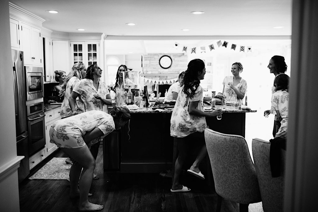Black and white photo of a bride and bridesmaids celebrating and laughing in a kitchen before getting ready for a wedding.
