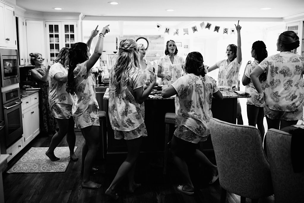 Black and white photo of a bride and bridesmaids celebrating and laughing in a kitchen before getting ready for a wedding.
