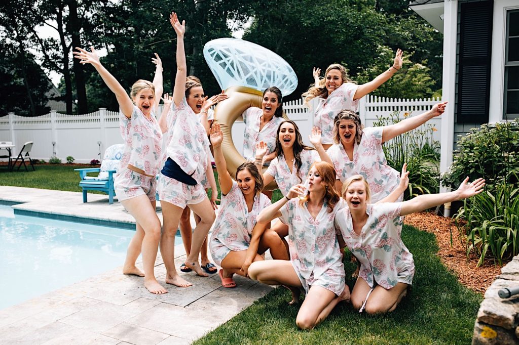 A bride and her bridesmaids celebrate next to a pool all wearing matching outfits before getting ready for the wedding day. The bride is holding a floatie that looks like a wedding ring.