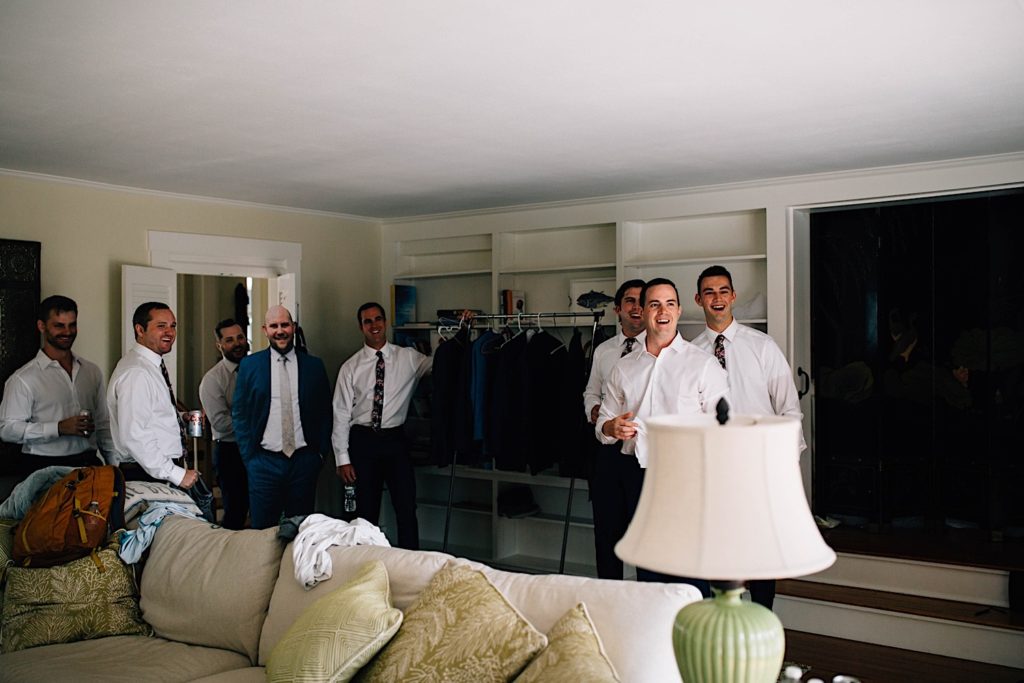 Groomsmen all dressed in button down shirts and ties stand and smile towards the groom who is to the right out of frame.