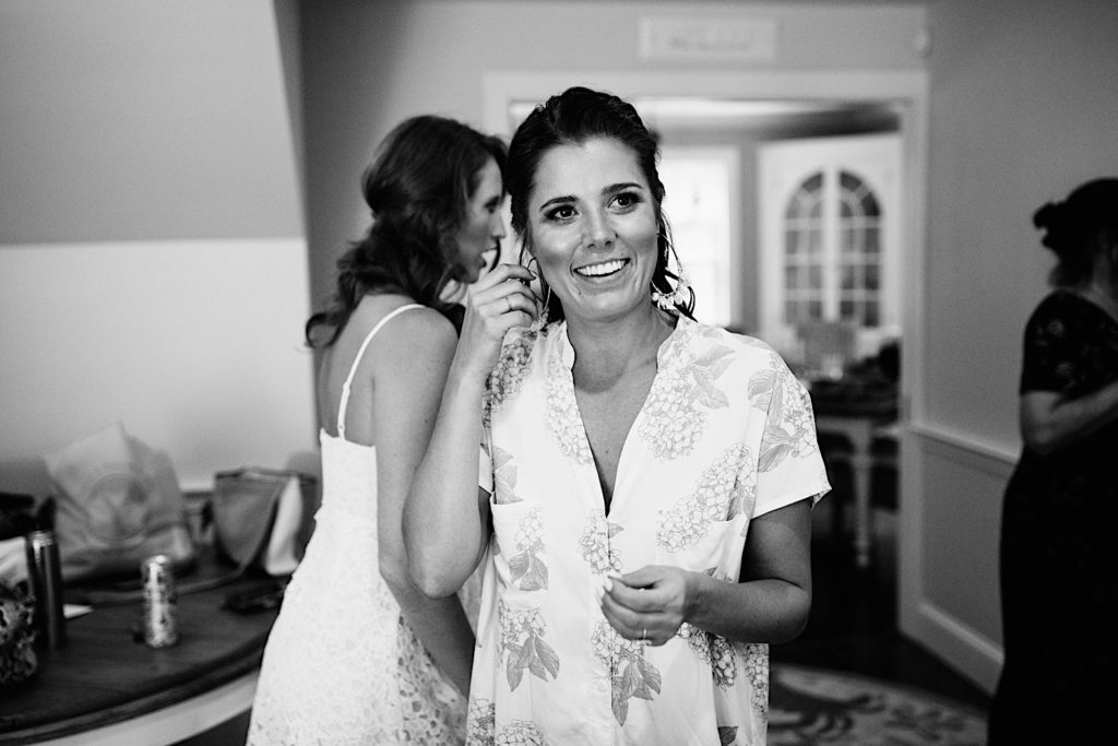 Black and white photo of a bride smiling as she touches one of her earrings with 2 bridesmaids in the background.