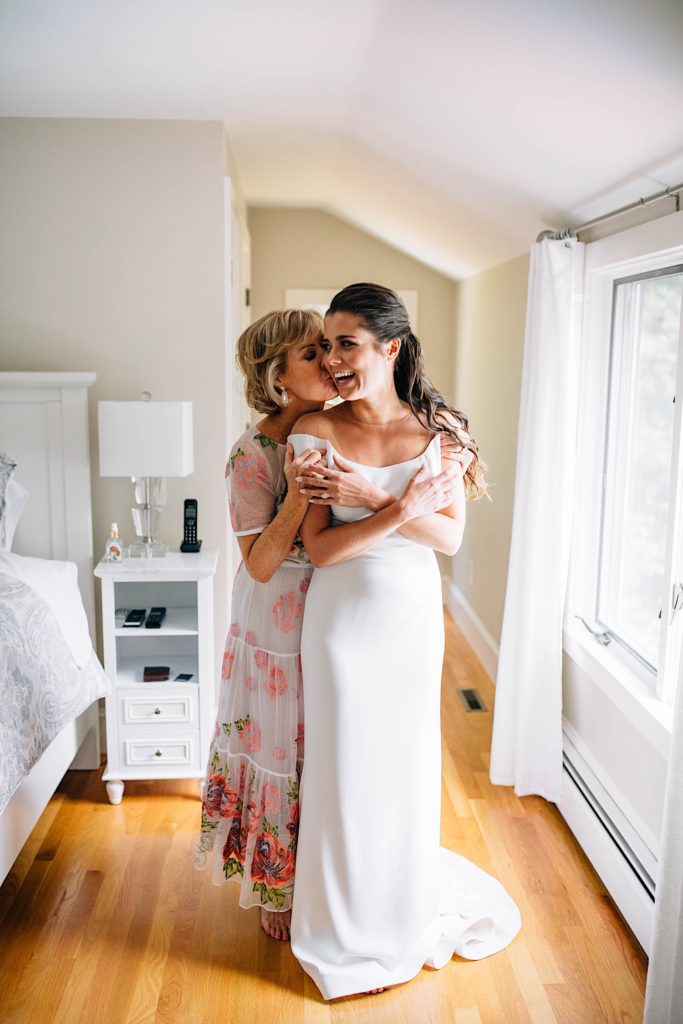 A bride smiles while wearing her wedding dress as her mother stands behind her and kisses her on the cheek.
